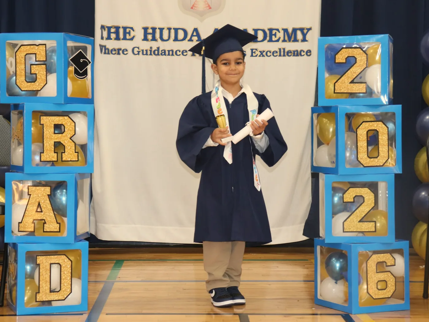 A kindergarten graduate in cap and gown holding a diploma in front of the school&rsquo;s banner